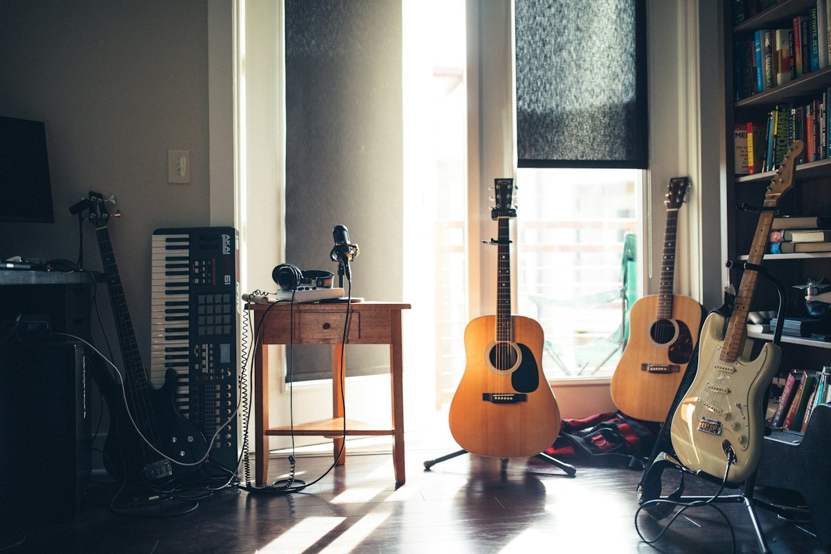 Acoustic and electric guitars on stands in a sunlit home music room with a keyboard and bookshelves