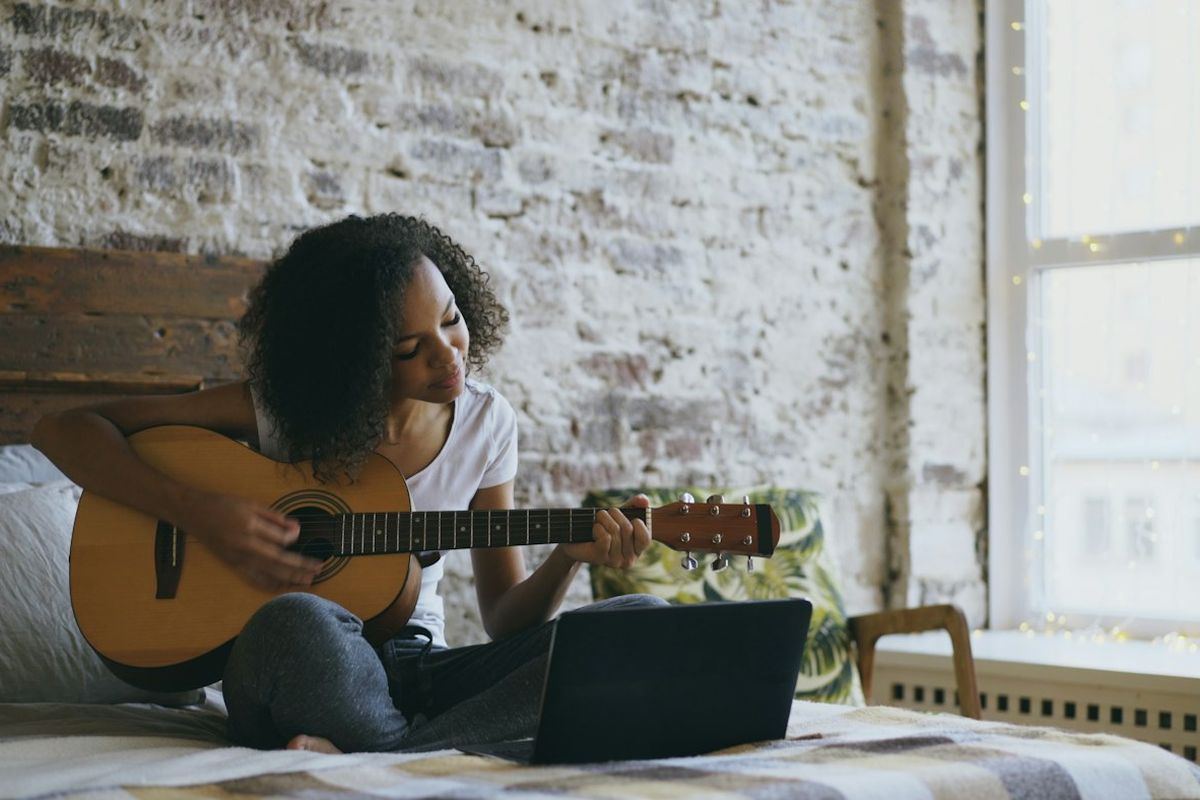 Young woman playing an acoustic guitar on her bed while following a lesson on a laptop