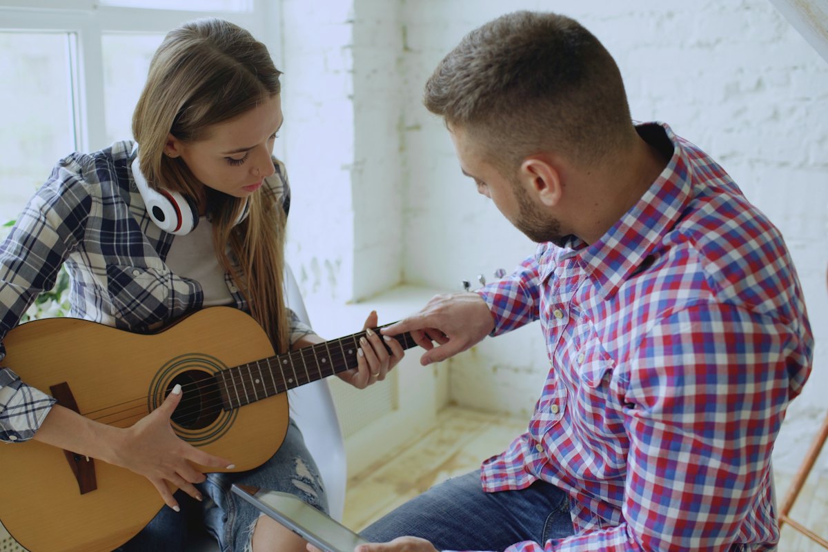 Guitar teacher pointing to the fretboard while a teenage student plays an acoustic guitar