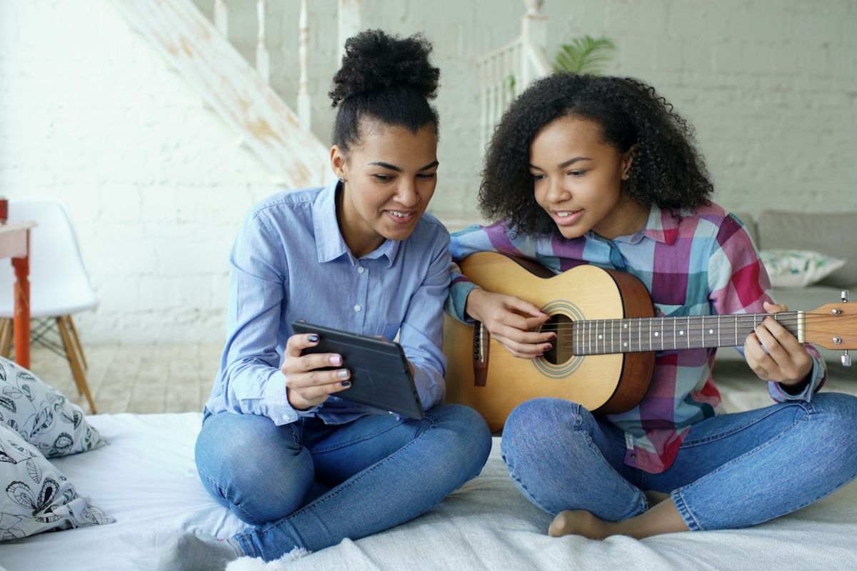 Two young women sitting on a bed, one holding a guitar, watching a lesson on a tablet