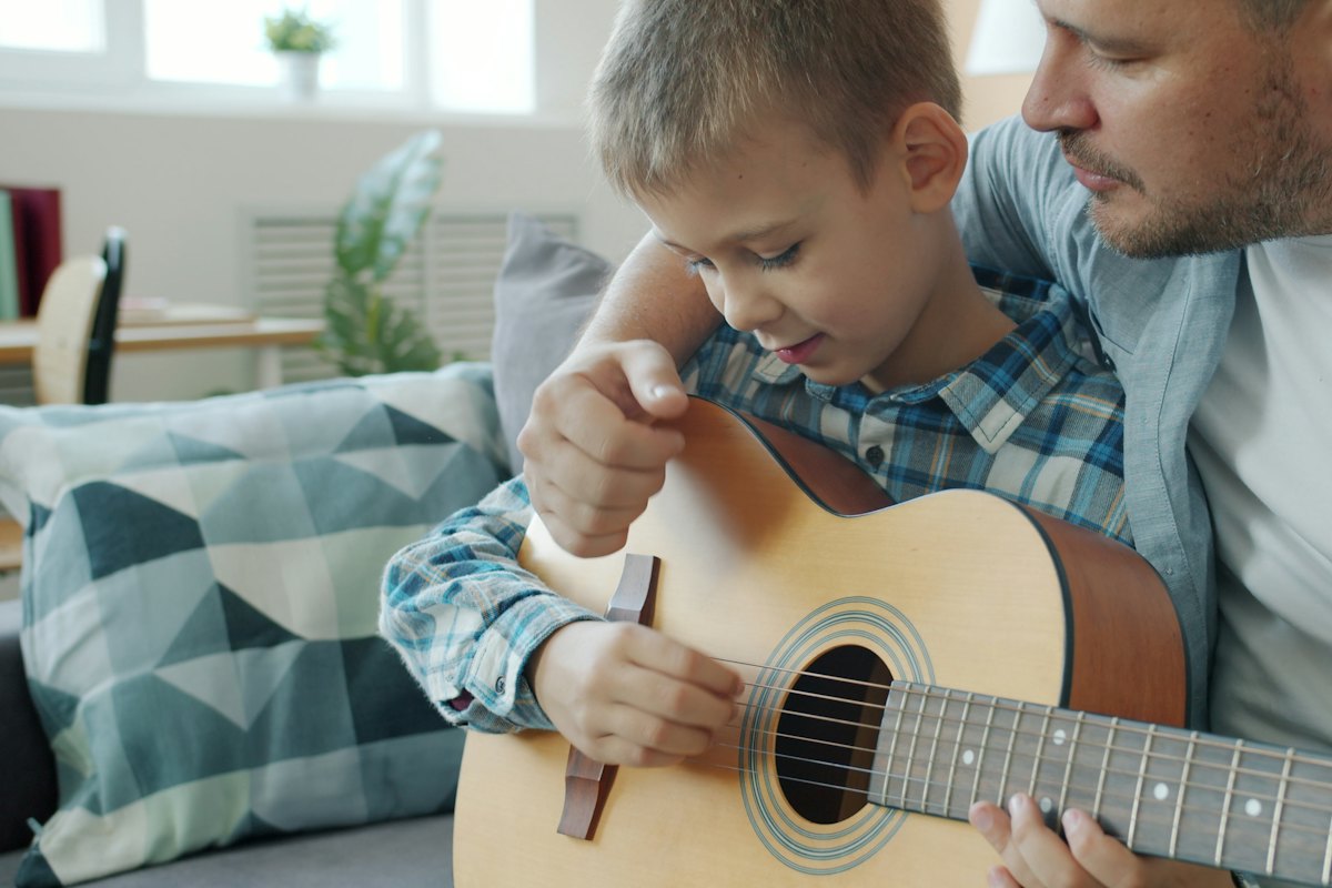 Father guiding his young son&rsquo;s hands on an acoustic guitar at home