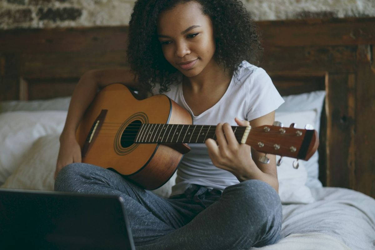 Young woman practising acoustic guitar on her bed with a laptop in front of her