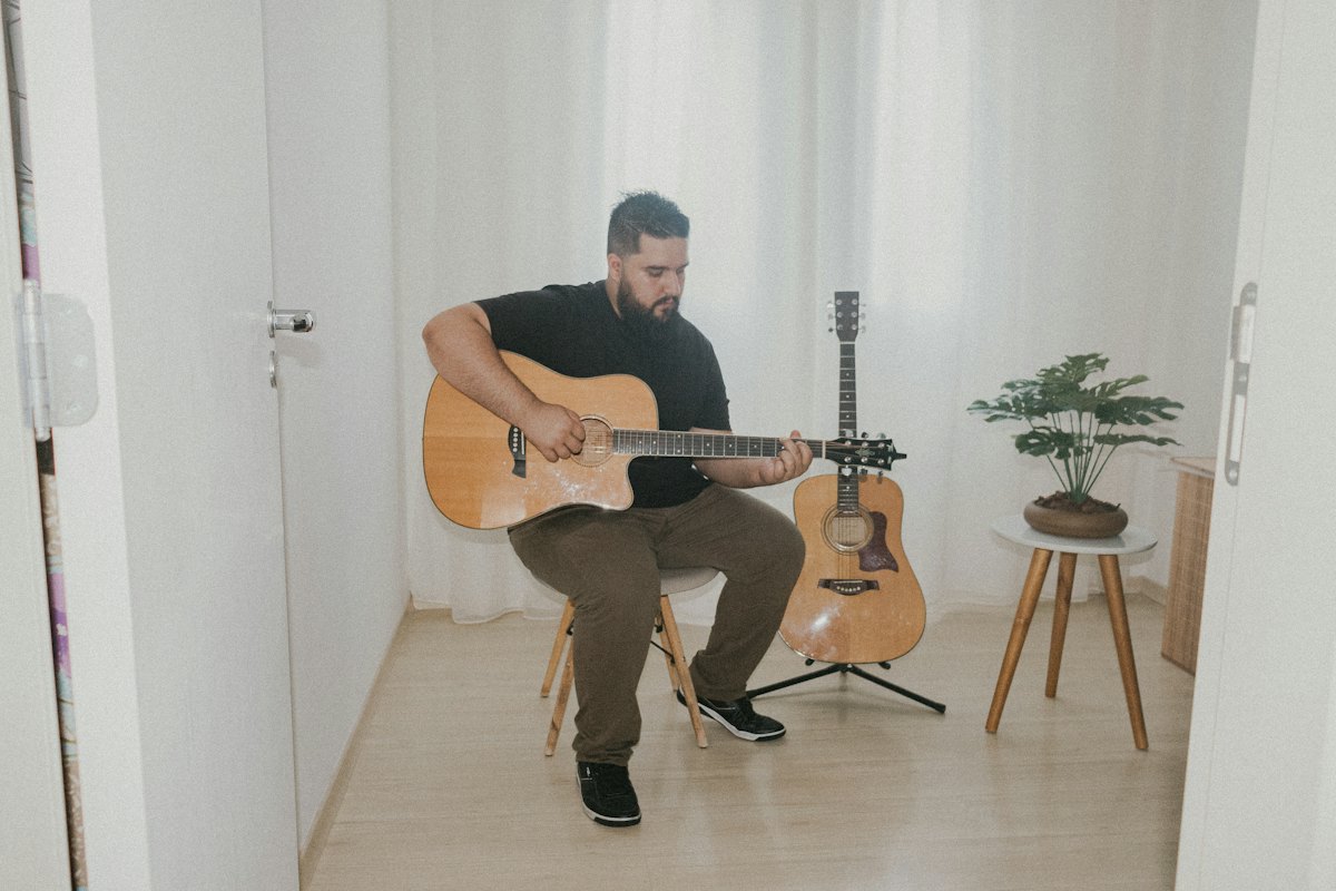 Man practising acoustic guitar alone on a stool in a bright, minimal room, with a second guitar on a stand