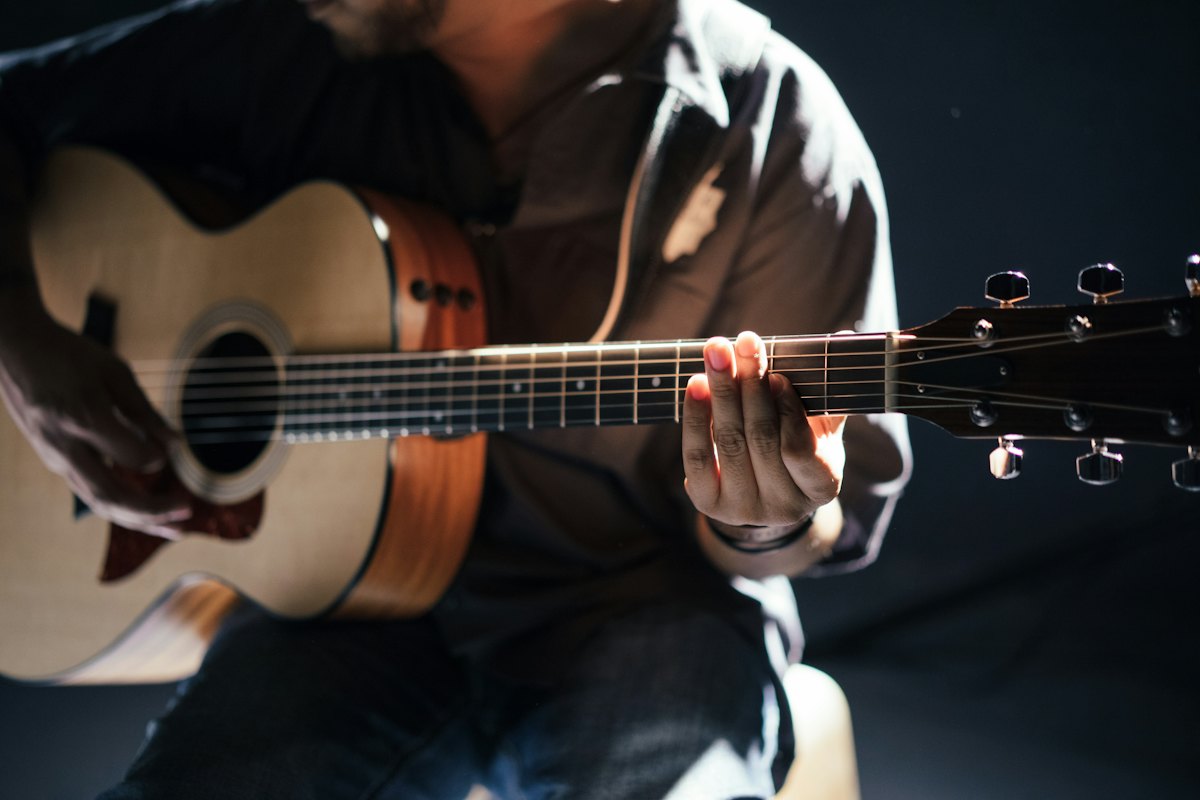 Man in a dark shirt fretting a chord on an acoustic guitar in low light