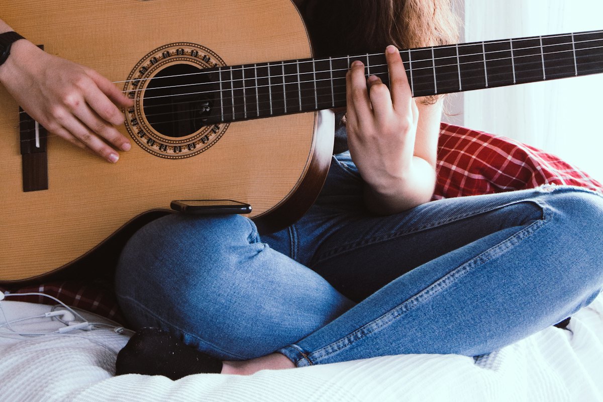 Person sitting cross-legged on a bed, practising an acoustic guitar alone