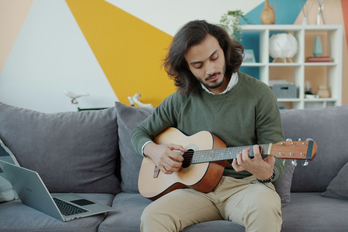 Man playing an acoustic guitar on a sofa with a laptop open beside him