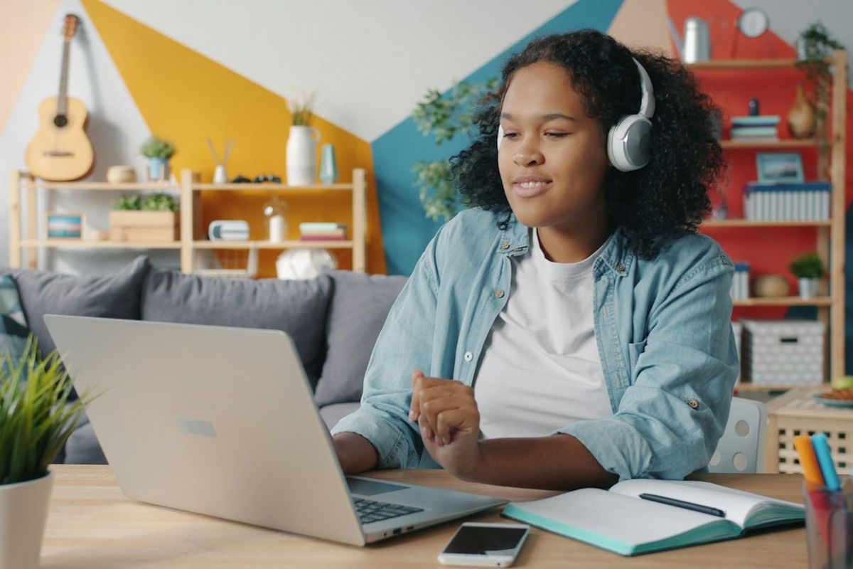 Young woman with headphones taking notes at a laptop, with guitars on the wall behind her