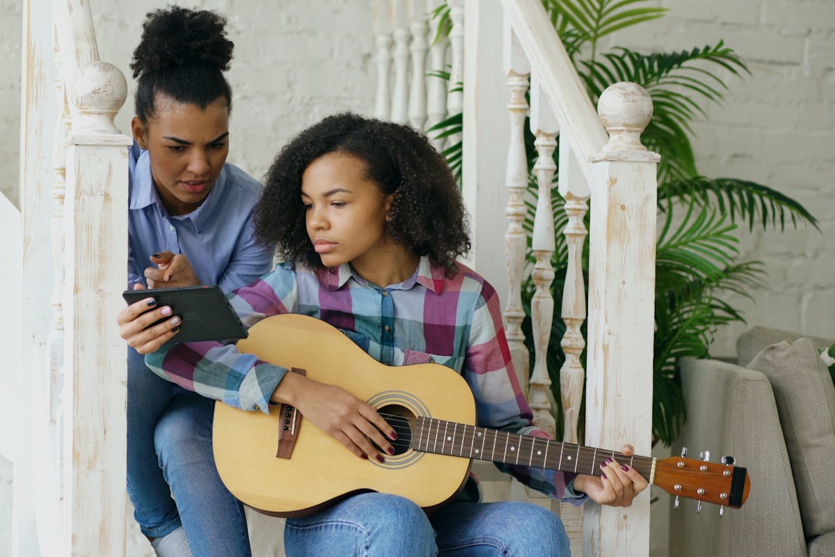 Two young women on a staircase following a guitar lesson on a tablet, one holding an acoustic guitar