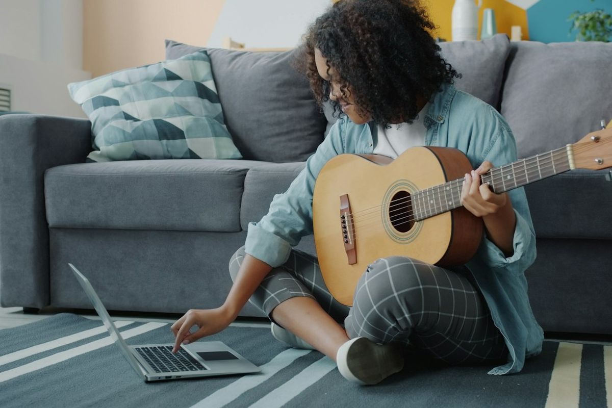 Woman sitting on the floor with an acoustic guitar, reaching for a laptop beside her